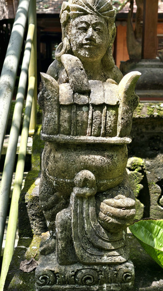 Stone statue of a robed figure with elaborate headgear in a mossy outdoor setting. Sunlight casts patterns on leaves and steps nearby.