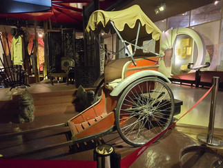 Historic rickshaw with beige canopy in museum; red rope barrier. Display includes antiques and "Guernica 1937" text on the wall.