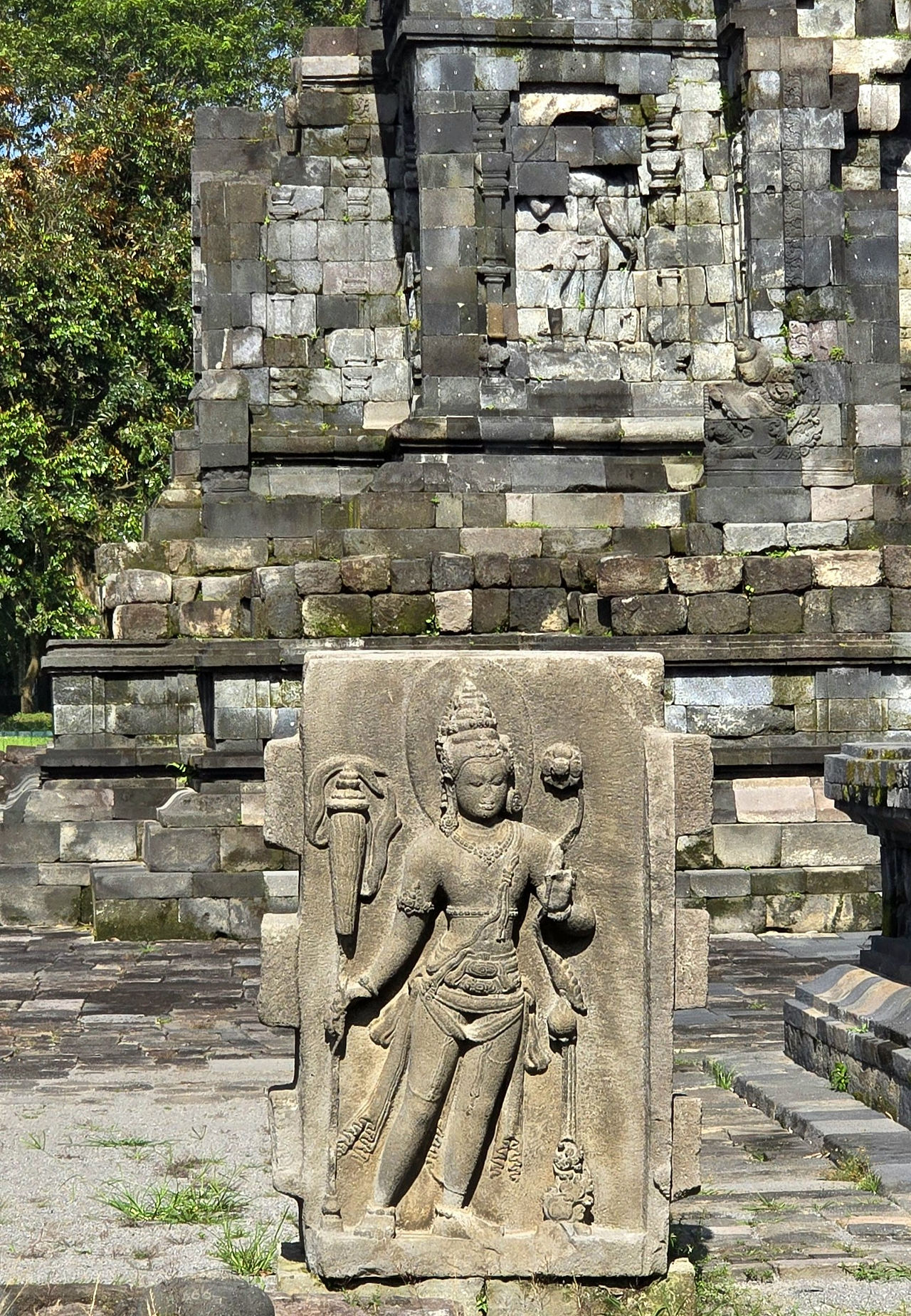 Ancient stone carving of a deity at a temple with intricate details, surrounded by lush greenery and stone ruins under clear daylight.