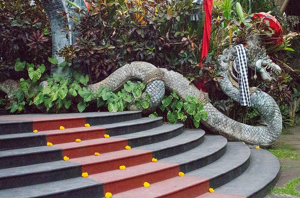 Stone dragon sculpture with intricate details beside curved steps adorned with scattered yellow flowers. Lush plants form the backdrop.