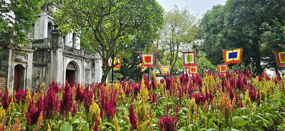 Colourful flags and vibrant red and yellow flowers in front of an ancient stone building, surrounded by lush green trees.