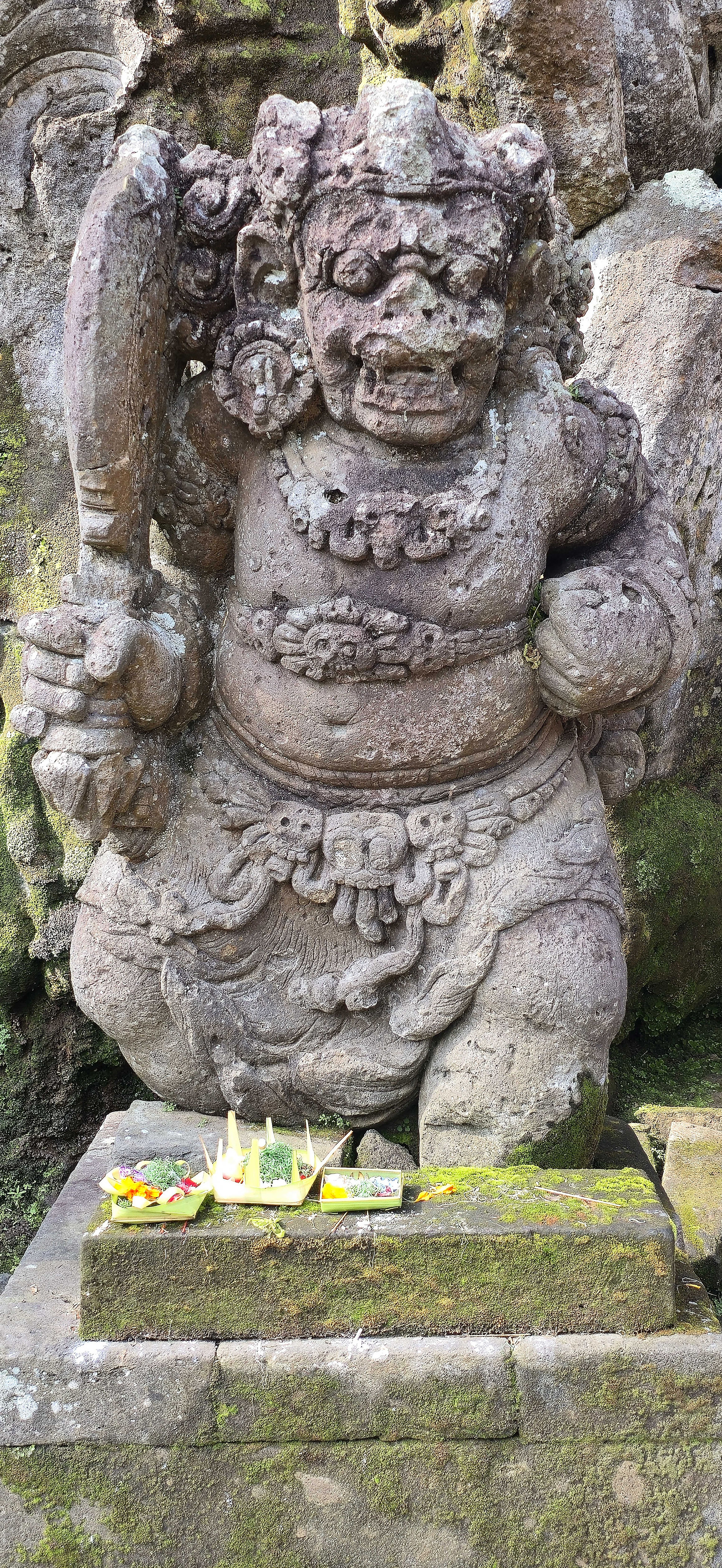 Stone statue of a fierce figure holding a club, surrounded by moss. Colourful offerings are placed at the base on a mossy stone platform.