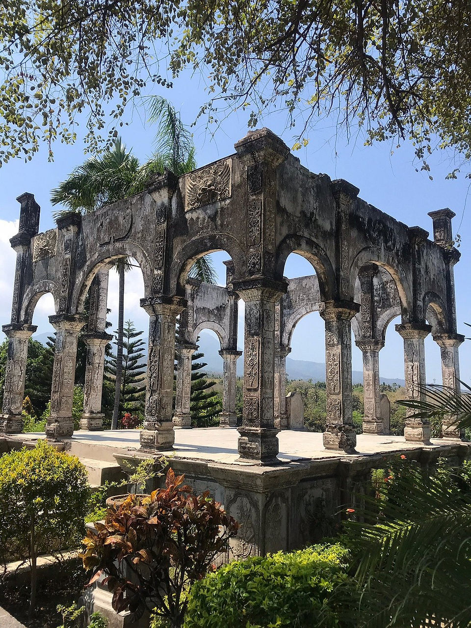 Stone pavilion with ornate arches, surrounded by lush greenery and palm trees under clear blue sky. Peaceful and historic atmosphere.