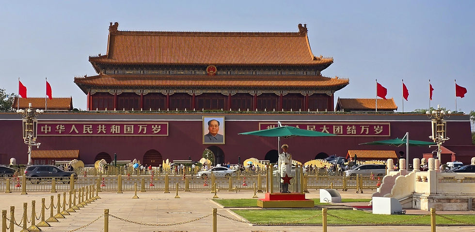 Red palace gates with large portrait, flanked by red flags. Guard stands under green umbrella. Chinese text on wall, cars and people visible.