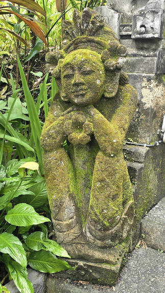 Moss-covered stone statue of a serene figure in a lush garden setting, surrounded by greenery, beside a mossy stone wall.