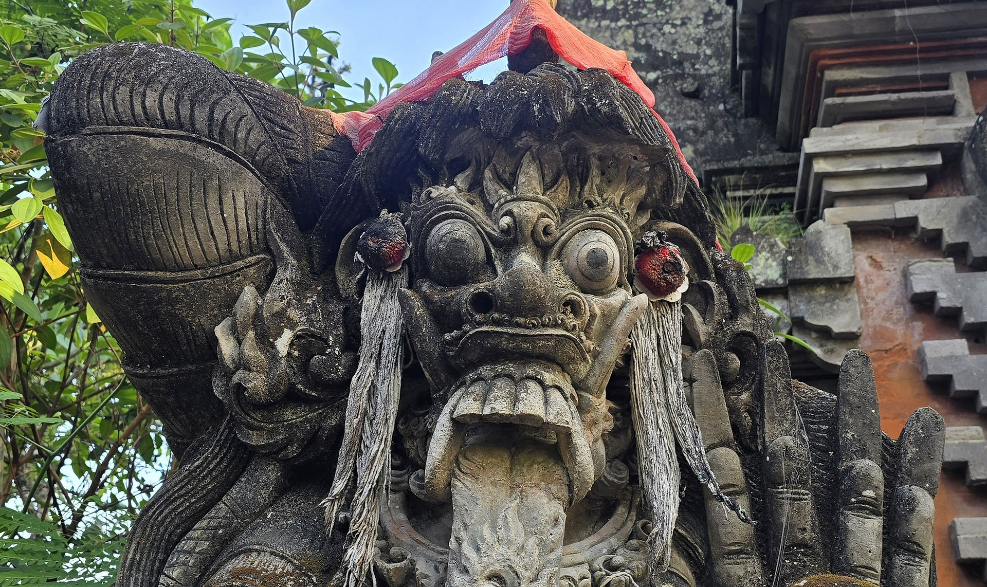 Weathered stone guardian statue with bulging eyes and red cloth, set among temple ruins and greenery under a bright sky