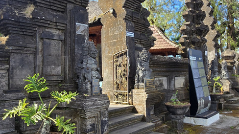 Stone temple gate with guardian statues, ornate metal gate, and informational signboards in a sunny tropical courtyard