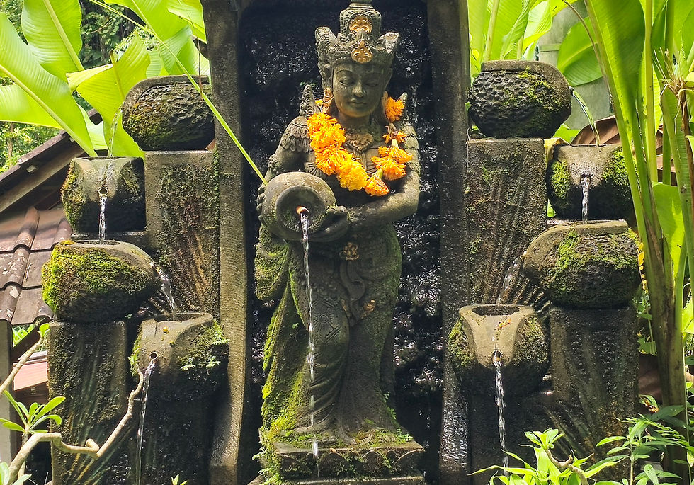 Ornate stone statue with orange marigold garland, water flowing from an urn, surrounded by lush green leaves and mossy stone pots.