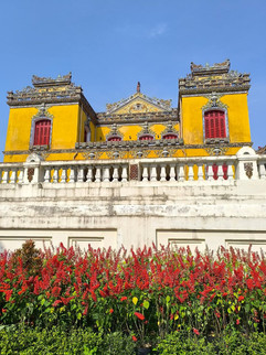 Yellow ornate temple with red windows, lined with vibrant red flowers, set against a clear blue sky. Elegant and historic atmosphere.