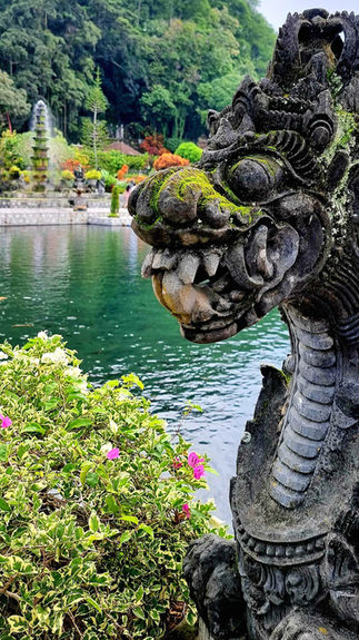 Stone dragon sculpture with moss by a pond, lush greenery and colorful flowers. Fountain and trees in background under a cloudy sky.