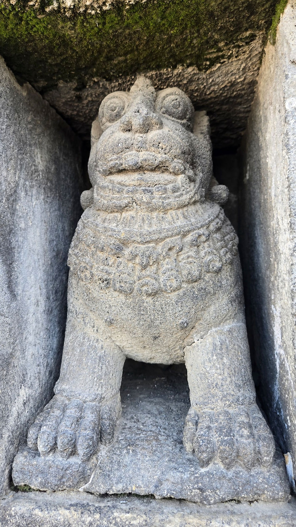 Stone lion statue in a niche with detailed carvings, surrounded by grey stone. Green moss above adds texture to the ancient setting.