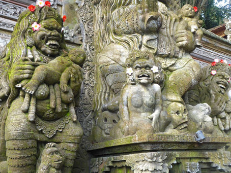 Ornate stone carvings of mythical figures holding babies, covered in moss, with flowers in hair, against detailed temple background.