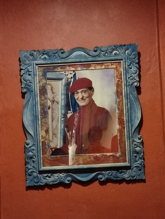 Portrait of a smiling Antonio Blanco in a red beret and jacket holds brushes, set in an ornate blue frame on a red textured wall.