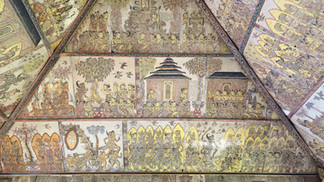 Detailed mural on a temple ceiling with scenes of deities, trees, and buildings. Vibrant reds, yellows, and intricate patterns evoke history.