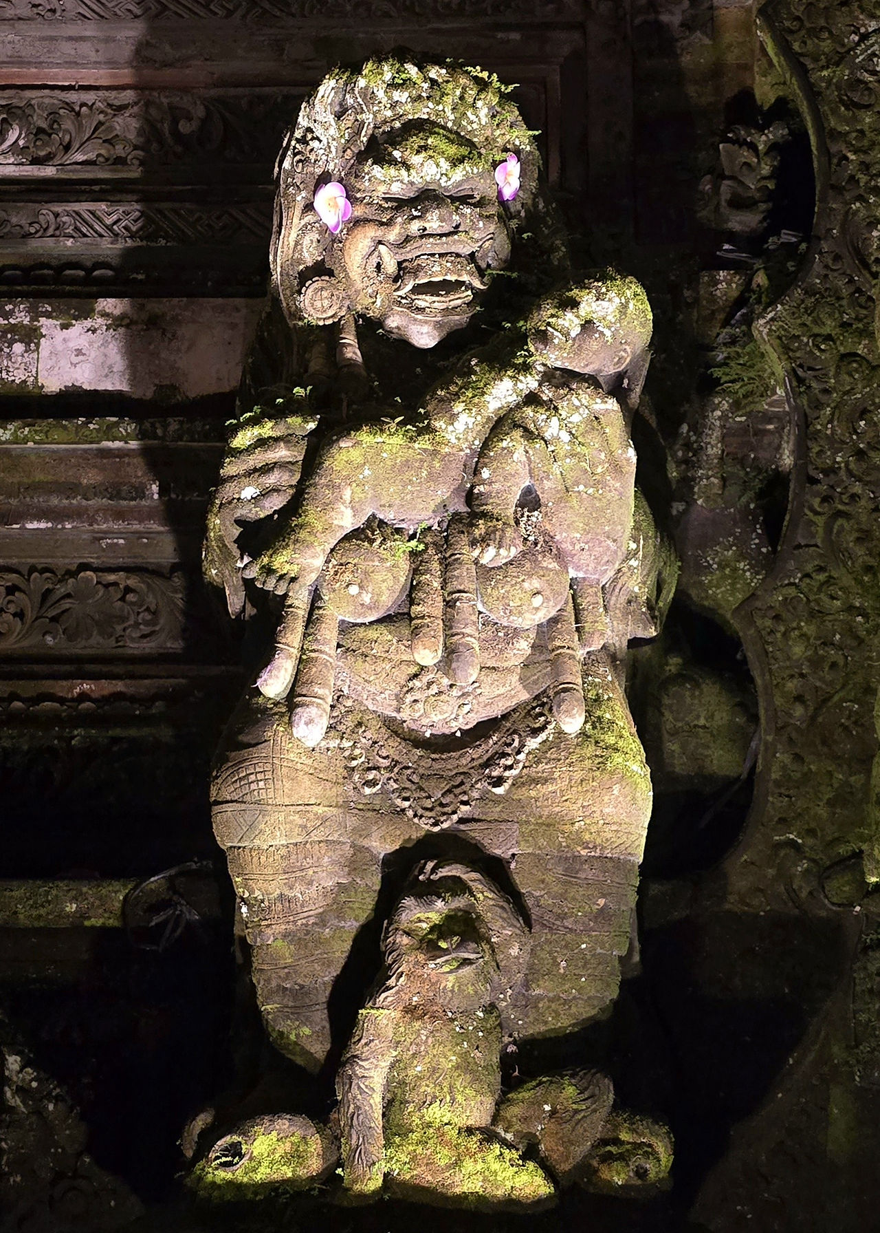 Stone statue of a mythical creature with intricate carvings, moss-covered, and adorned with pink flowers. Set against an ornate backdrop.