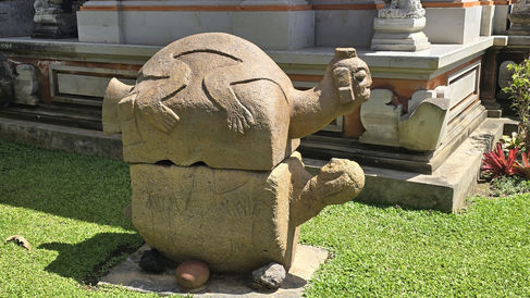 Stone sculpture of a rounded figure with outstretched arms and animal carvings, set on grass near a traditional architectural structure.