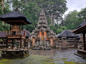 Ancient temple courtyard with ornate stone carvings and statues, surrounded by large trees. Overcast sky creates a serene, mystical ambiance.