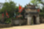 Ancient stone gate with red and yellow triangular flags, lush greenery in background, under a sunny blue sky. Peaceful and historical setting.