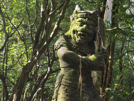 Moss-covered stone statue with a fierce expression in a lush, green forest. Sunlight filters through the leafy branches, creating a serene scene.