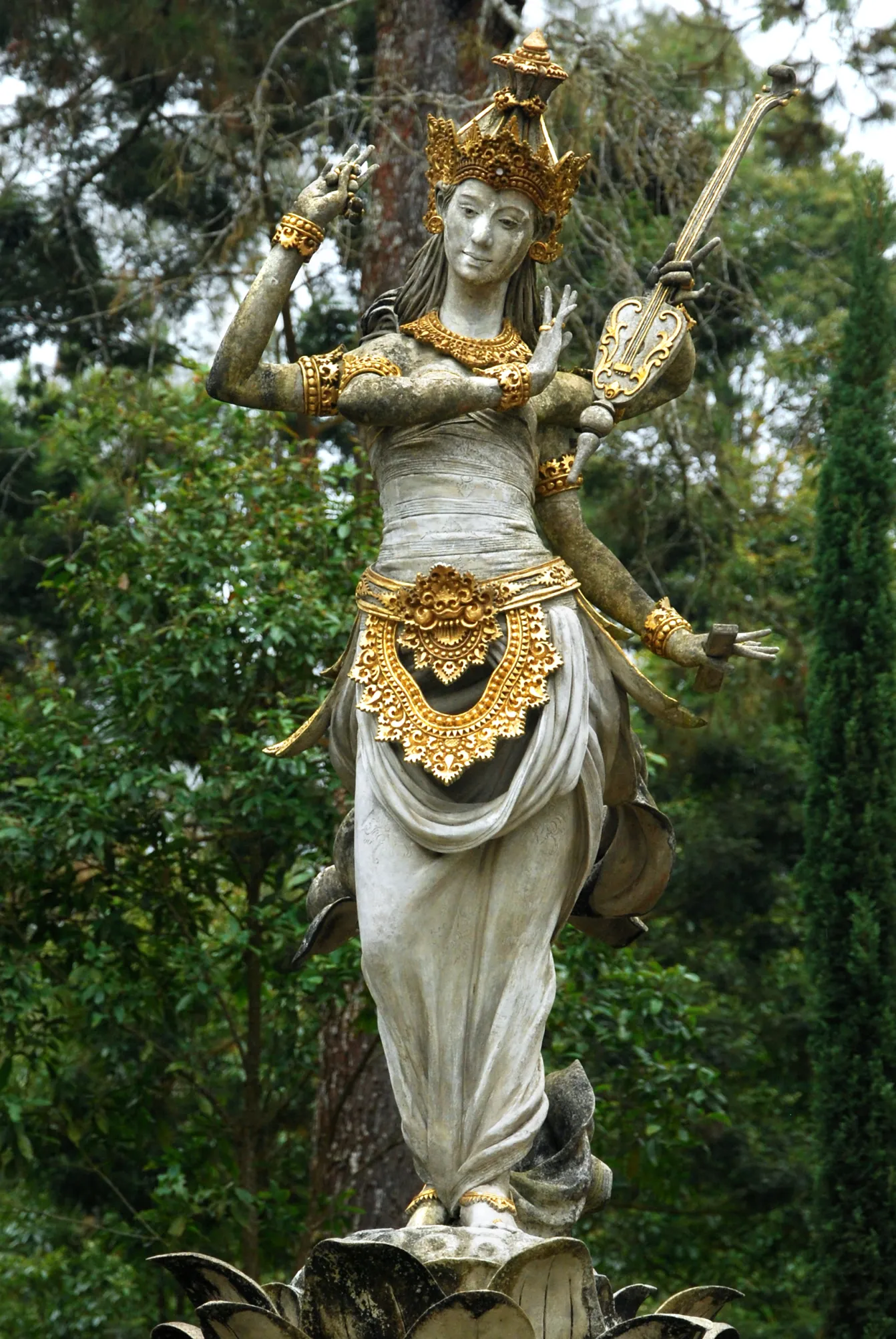 Statue of Saraswati with golden accents, poised elegantly with upraised arms. Set in a lush green forest, evoking serenity and grace.