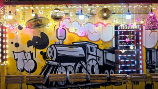 Graffiti train on a vibrant yellow wall with colourful string lights and pink and yellow bulbs, creating a lively, festive atmosphere.