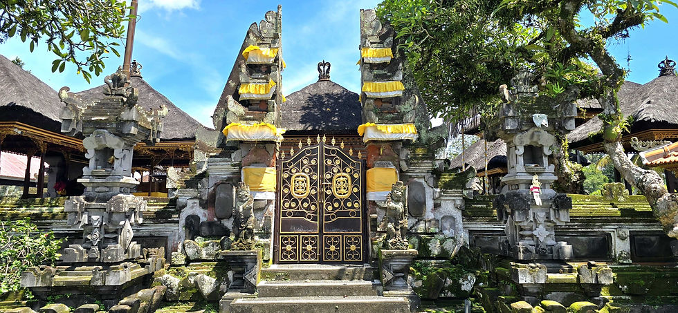 Ornate temple gate with stone carvings, yellow cloths, and intricate ironwork. Thatched roofs and greenery in the background, under blue sky.