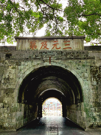 Ancient Stone archway with Chinese characters, framed by lush green leaves. Sunlight filters through, illuminating a bustling street beyond.