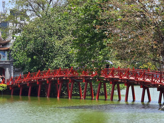 Red wooden bridge over a green lake, surrounded by lush trees. People walk on the bridge, leading to a traditional temple gate.