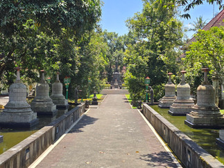 Pathway flanked by stone stupas and water features, surrounded by lush trees and buildings with red-tiled roofs under a clear blue sky.