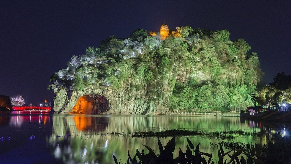 Night view of a lit elephant trunk hill with trees and a pagoda. A bright, glowing cave is reflected in a calm lake. Red bridge lights in the background.