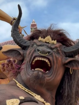 A fierce creature with horns and open mouth is adorned with gold, amid a dynamic scene of swords and warriors against a cloudy sky.