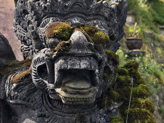 Moss-covered stone guardian statue with fierce carved face in a lush garden, weathered black-grey texture