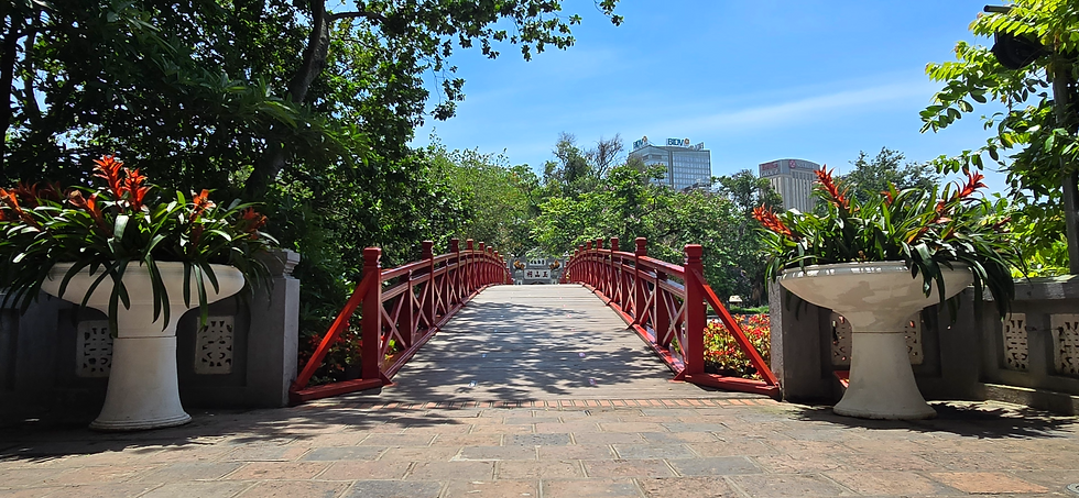 Red bridge with white railings, flanked by large potted plants with red flowers. Lush green trees surround the scene under a clear blue sky.