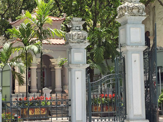 Ornate gate to a garden with lush palms, red flowers, and a pale pink building under a sunny blue sky. Signs with text in the foreground.