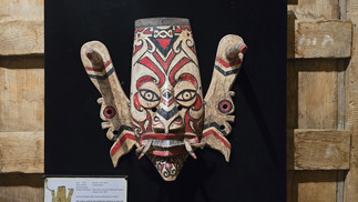 Wooden tribal mask with red and black patterns on a black background. Board underneath shares details in multiple languages. Rustic wooden frame.