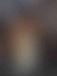 Performer in a colourful costume and mask stands in the center, surrounded by seated shirtless men in a crowded, engaged audience setting.