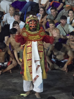 Performer in a colourful costume and mask stands in the center, surrounded by seated shirtless men in a crowded, engaged audience setting.