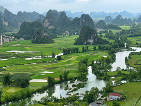 Lush green landscape with rice paddies, a winding river, and karst mountains in the background. Scattered houses with red roofs. Serene mood.