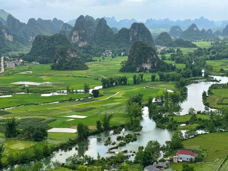 Lush green landscape with rice paddies, a winding river, and karst mountains in the background. Scattered houses with red roofs. Serene mood.
