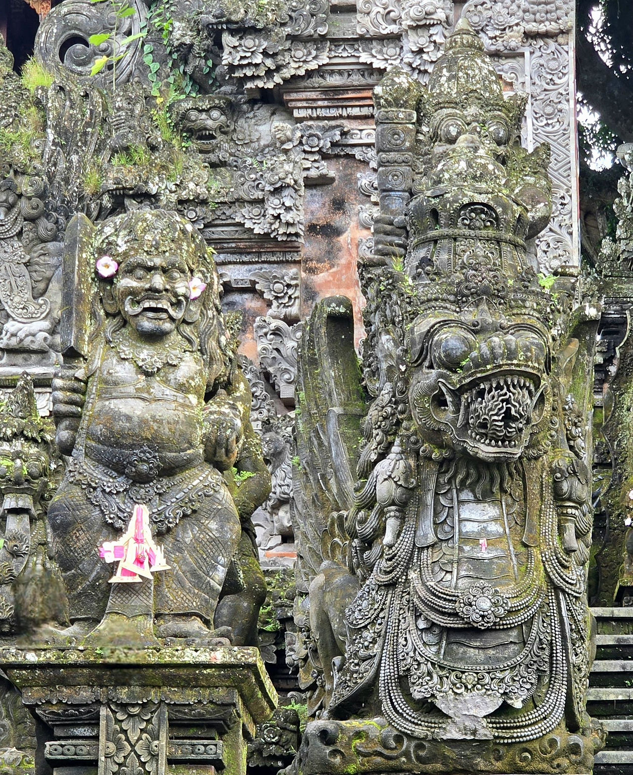 Mossy stone naga and rangda statues adorned with pink flowers, in a dimly lit area with intricate carvings on the wall, creating a mystical atmosphere.