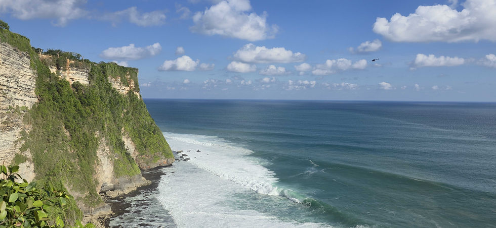 Cliffside view of ocean waves hitting the shore beneath a blue sky with scattered clouds. A helicopter flies above the sea.