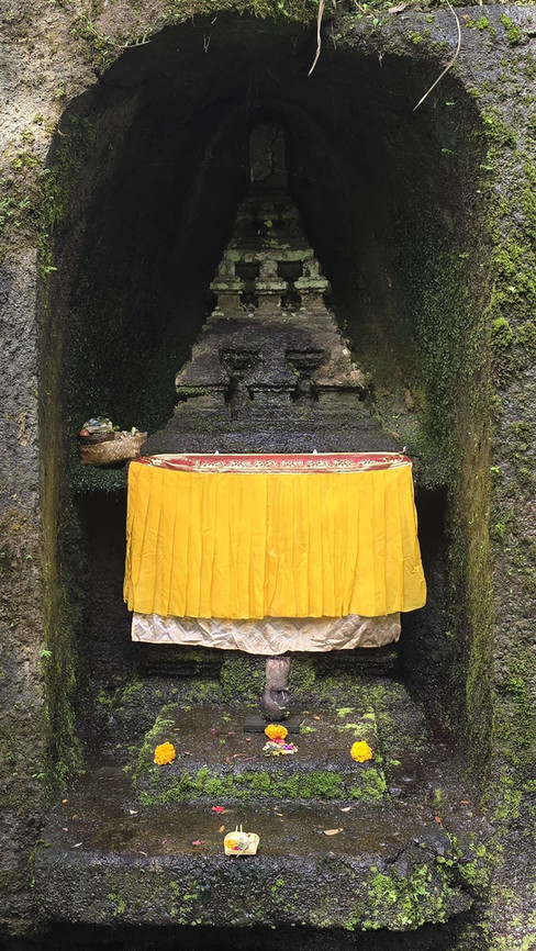 Stone shrine in a mossy cave, adorned with a yellow cloth. Offerings and marigold flowers are placed on the steps below.