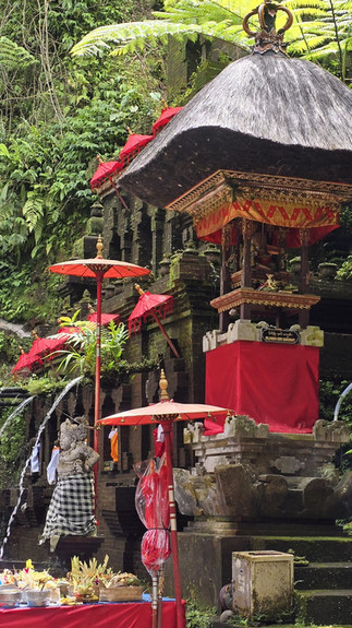 Balinese temple shrine with red parasols and offerings. Lush greenery surrounds the stone structure, creating a serene, sacred atmosphere.