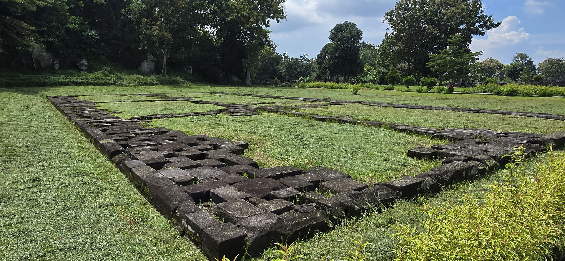 Ancient stone ruins on grassy field, surrounded by lush trees under a partly cloudy sky. The scene is peaceful and serene.