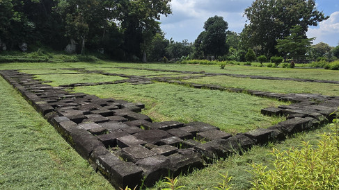Ancient stone ruins on grassy field, surrounded by lush trees under a partly cloudy sky. The scene is peaceful and serene.