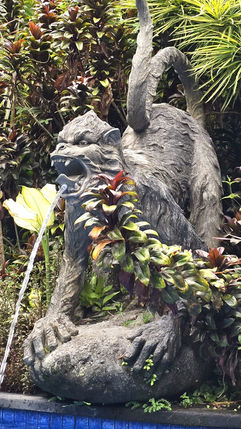 Stone monkey statue with open mouth, water flowing from it into a pool. Surrounded by lush green plants. Tranquil garden setting.