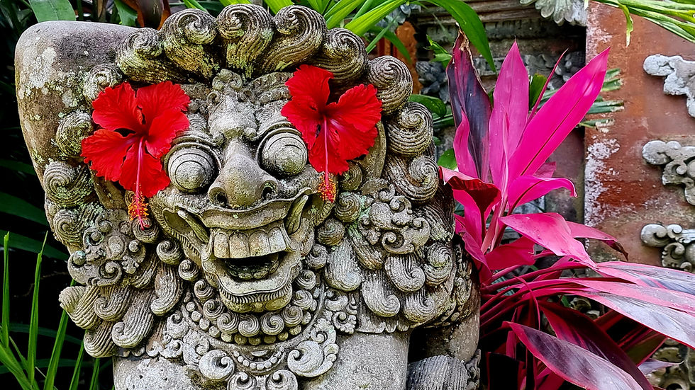 Stone statue with red hibiscus flowers as eyes, surrounded by lush green and pink foliage. Weathered appearance, smiling expression.