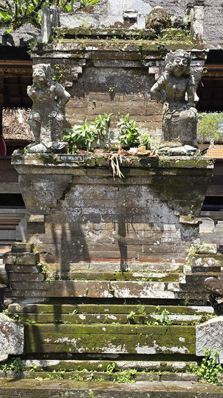 Ancient stone shrine with two statues, lush greenery, and "RATU TIRTA EMPUL" plaque. Sunlit, casting shadows on the mossy structure.