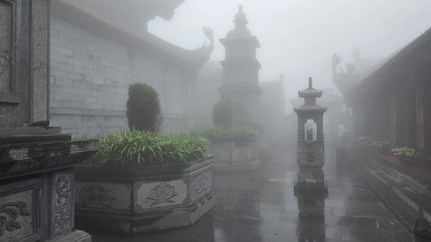 Foggy stone courtyard with ornate pillars, lush greenery, and Asian architectural details creates a mysterious, serene atmosphere.