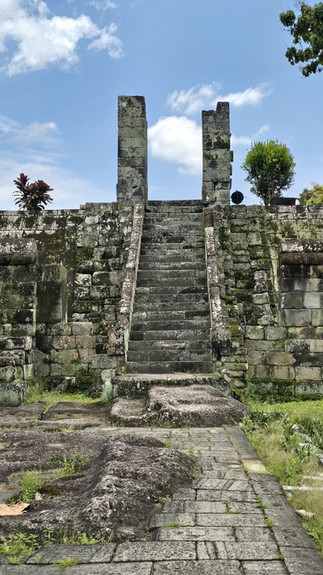 Ancient stone stairway with moss leads to a ruin under a bright blue sky. Greenery and scattered plants grow between stones.